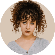 Portrait of a woman with curly hair and bangs wearing a gray top, against a beige background.